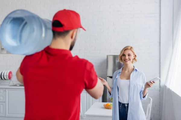 Back view of delivery man with big bottle of water near happy woman showing wow gesture, blurred foreground — Stock Photo