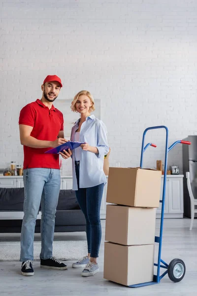 Happy arabian postman and blonde woman looking at camera near trolley with parcels — Stock Photo