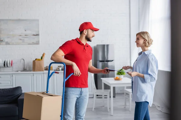 Muslim postman giving clipboard to woman while standing near trolley with parcels on blurred foreground — Stock Photo