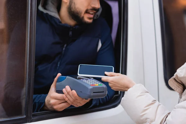 Partial view of woman holding smartphone near postman with payment terminal on blurred background — Stock Photo