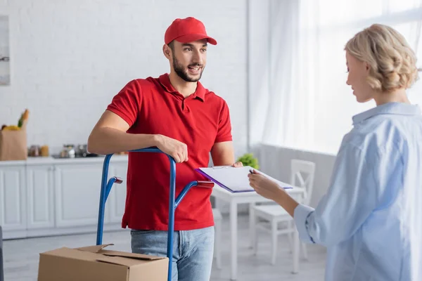 Blonde woman signing order on clipboard near muslim postman, blurred foreground — Stock Photo
