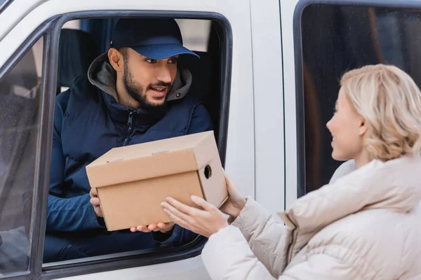 Musulman facteur dans camion donnant colis à femme blonde sur flou premier plan — Photo de stock