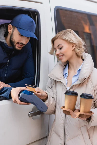 Donna sorridente con caffè per andare a pagare l'ordine con carta di credito vicino uomo consegna araba — Foto stock