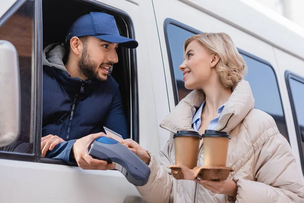 Arabian delivery man holding payment terminal near blonde woman with credit card and coffee to go — Stock Photo
