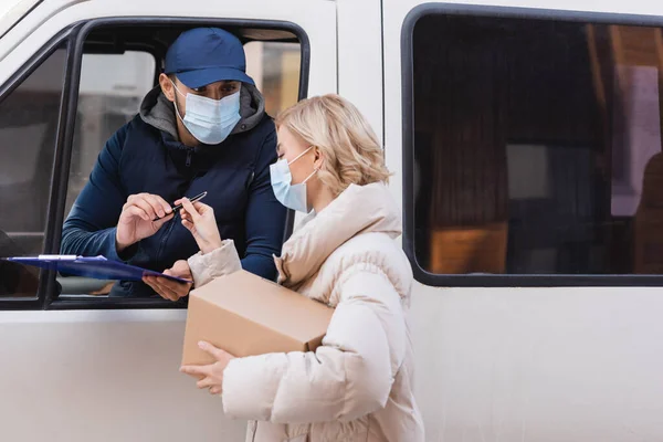 Muslim courier in medical mask giving pen to woman holding parcel — Stock Photo