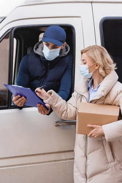 Blonde woman signing order on clipboard near arabian delivery man in medical mask — Stock Photo