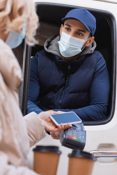 Uomo musulmano di consegna in maschera medica con terminale di pagamento vicino alla donna con smartphone in primo piano sfocato — Foto stock