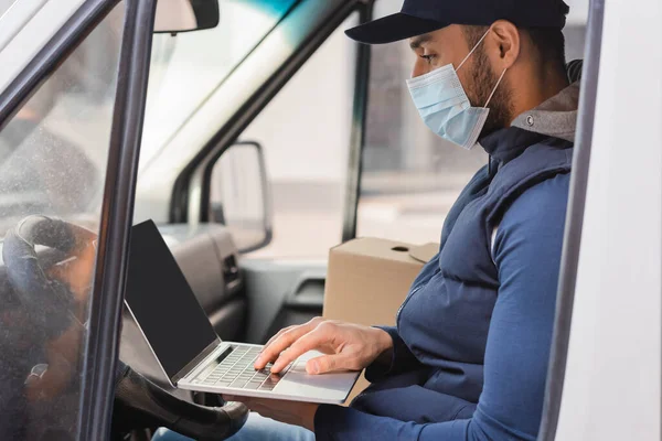 Arabe livreur en masque médical tapant sur ordinateur portable tout en étant assis dans la voiture — Photo de stock