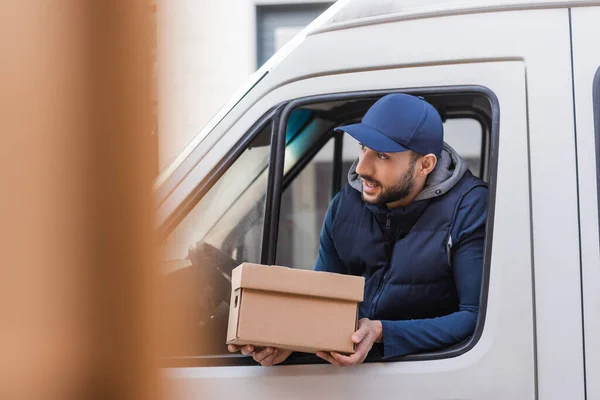 Muslim delivery man with carton box looking out car window on blurred foreground — Stock Photo