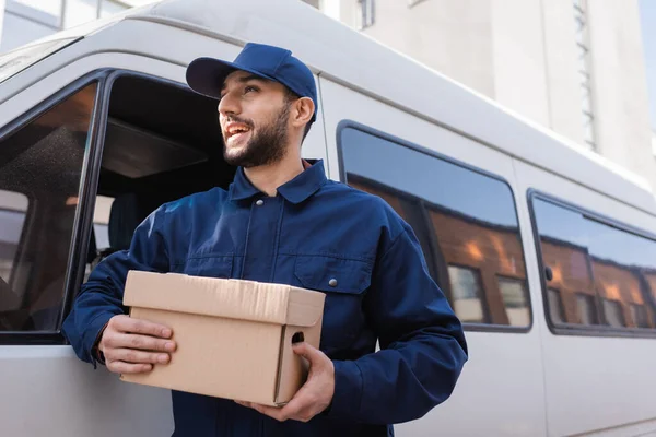 Happy arabian postman looking away while standing with parcel near car — Stock Photo