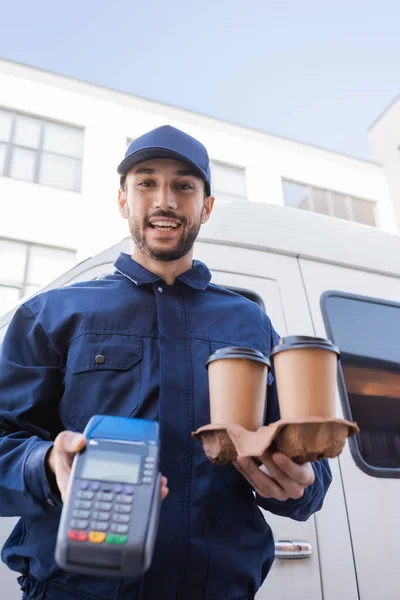 Heureux homme de livraison musulman tenant des tasses en papier et terminal de paiement sur le premier plan flou — Photo de stock