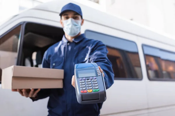 Mensajero árabe en máscara médica con terminal de pago y caja de cartón sobre fondo borroso — Stock Photo