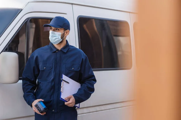 Muslim delivery man in medical mask looking away while standing near car on blurred foreground — Stock Photo