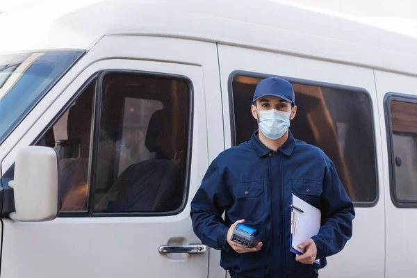 Jeune messager arabe en masque médical debout avec presse-papiers et lecteur de carte de crédit près de la voiture — Photo de stock