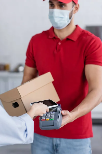 Arabe livreur en masque médical tenant terminal de paiement près de la femme avec smartphone, fond flou — Photo de stock