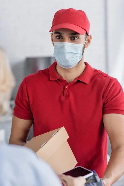 Muslim courier in protective mask holding payment terminal near woman with mobile phone on blurred foreground — Stock Photo