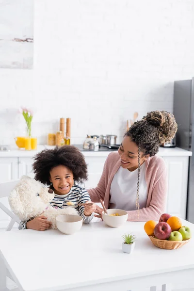 Cheerful african american girl holding spoon with corn flakes near teddy bear and smiling mom — Stock Photo