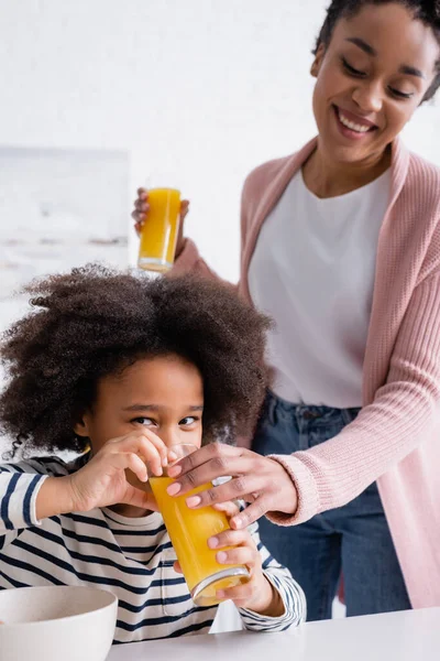 Africano americano niño bebiendo jugo de naranja de vidrio en la mano de la madre sobre fondo borroso - foto de stock
