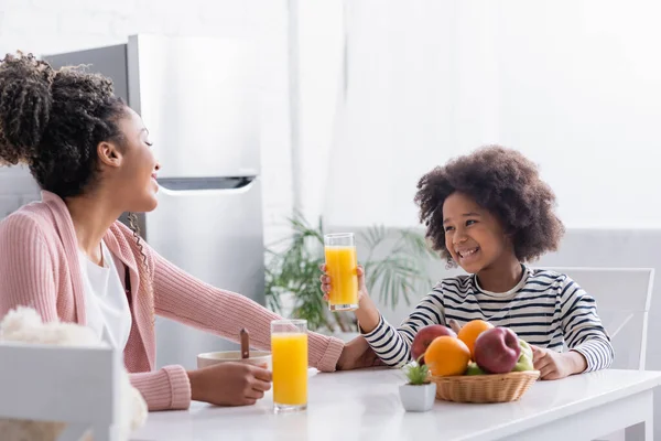 Alegre afroamericana chica sosteniendo vaso de jugo de naranja cerca de madre durante el desayuno - foto de stock
