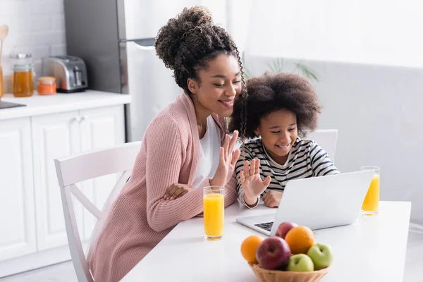 Bambino afroamericano allegro con madre che agita le mani durante la videochiamata sul computer portatile in cucina — Foto stock