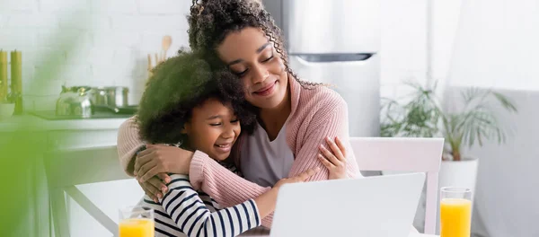 Happy african american mom and daughter embracing while watching film on laptop, blurred foreground, banner — Stock Photo