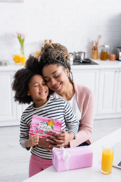 Feliz africana americana mujer abrazando hija celebración feliz madres día tarjeta cerca de caja de regalo - foto de stock