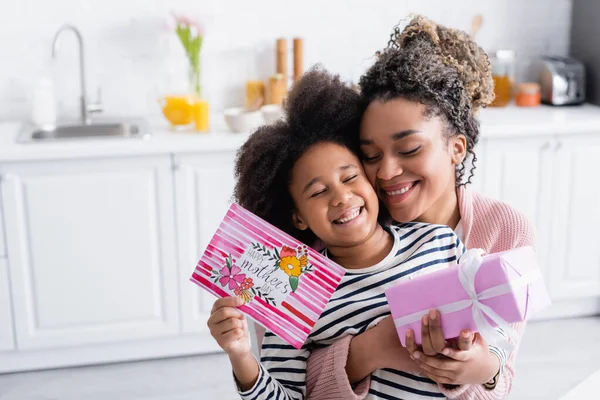 Mujer afroamericana emocionada abrazando a hija feliz sosteniendo caja de regalo y tarjeta de día de madres felices - foto de stock