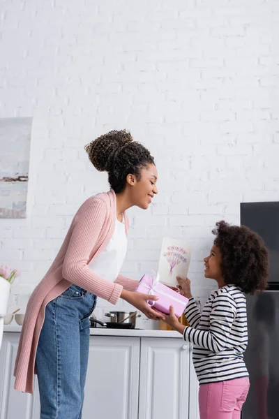 Vista lateral del niño afroamericano presentando regalo y tarjeta de felicitación a mamá feliz en la cocina - foto de stock