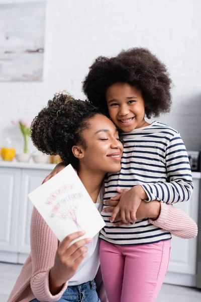 Alegre africana americana mujer con los ojos cerrados celebración feliz madres día tarjeta mientras abrazo sonriente hija - foto de stock