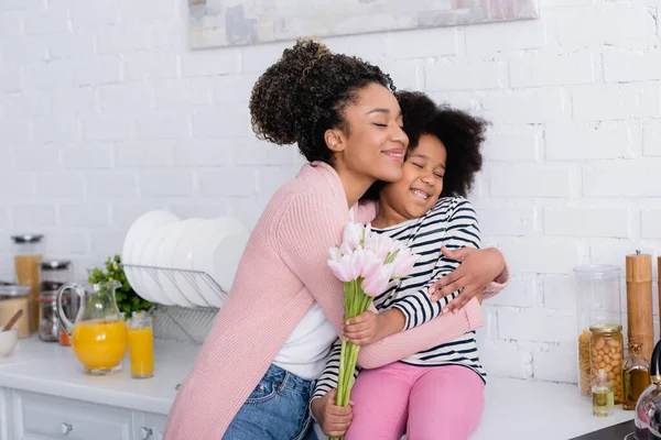 Pleased african american woman embracing daughter sitting on kitchen counter with tulips — Stock Photo