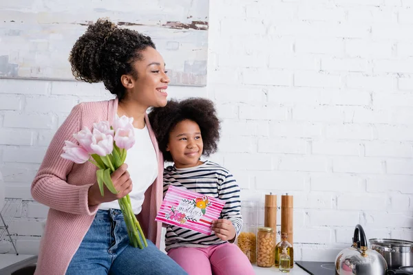 Glückliche afrikanisch-amerikanische Frau und Mädchen mit glücklicher Muttertagskarte lächelnd und wegschauend in der Küche — Stockfoto