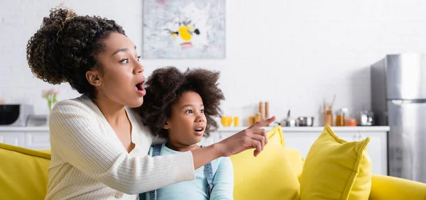 Astonished african american woman looking away and pointing with finger near daughter at home, banner — Stock Photo