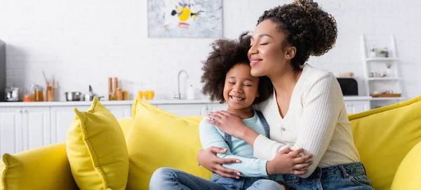 Niño afroamericano feliz y mamá abrazando con los ojos cerrados en el sofá en casa, pancarta - foto de stock