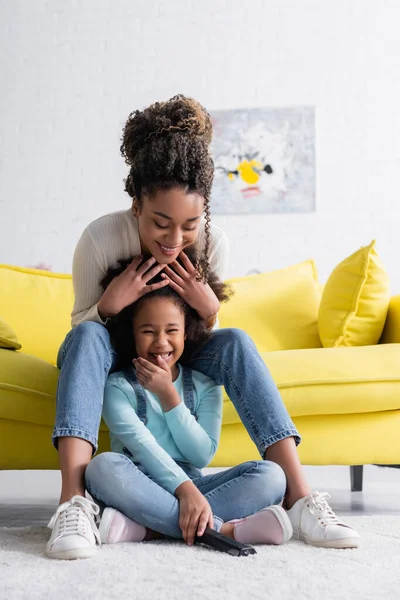 Laughing african american child covering mouth with hand while watching comedy on floor near mother — Stock Photo