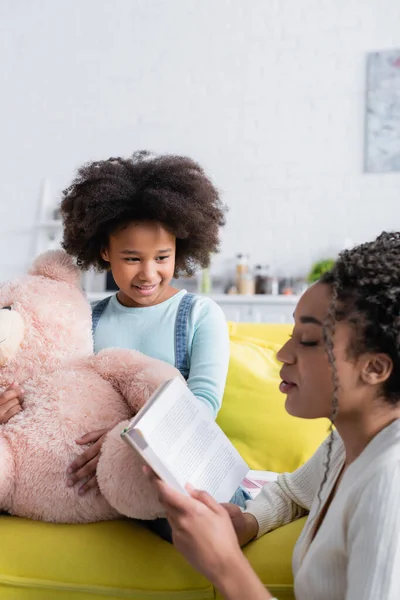 African american woman reading book to daughter sitting on sofa with teddy bear — Stock Photo