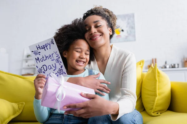 Pleased african american woman holding gift box and embracing daughter with happy mothers day card — Stock Photo