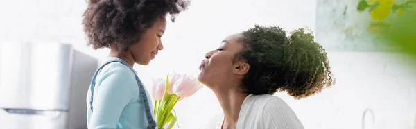 Side view of african american woman blowing air kiss to smiling daughter with fresh tulips, banner — Stock Photo