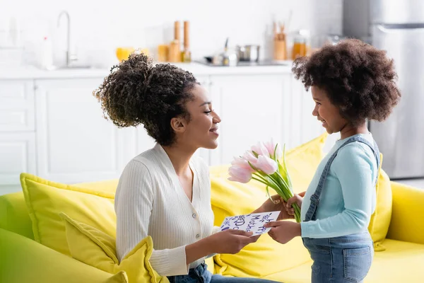 Smiling african american child presenting tulips and happy mother day card to happy mom — Stock Photo