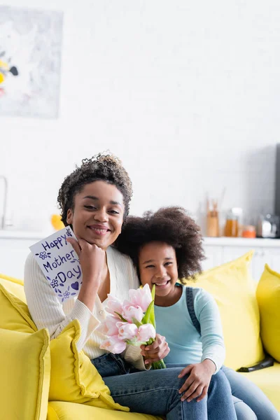 Alegre africana americana chica sentado cerca feliz mamá celebración tulipanes y feliz madres día tarjeta - foto de stock