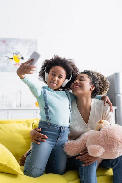African american girl in headphones and woman with teddy bear taking selfie while sticking out tongues — Stock Photo