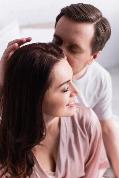 Мужчина целует улыбающуюся жену с закрытыми глазами в спальне — стоковое фото
