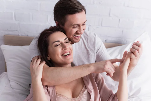 Smiling man pointing at palm of cheerful wife on bed — Stock Photo