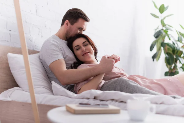 Hombre besar esposa cerca de teléfono inteligente y taza en primer plano borrosa en el dormitorio - foto de stock