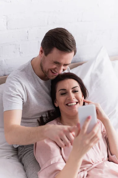 Smiling adult couple using smartphone on blurred foreground on bed — Stock Photo