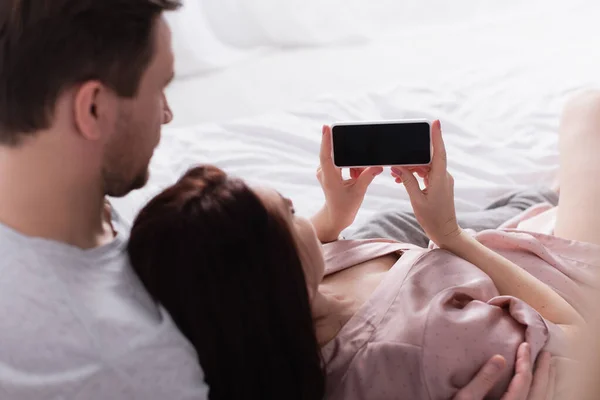 Smartphone with blank screen in hands of woman lying near husband on bed — Stock Photo