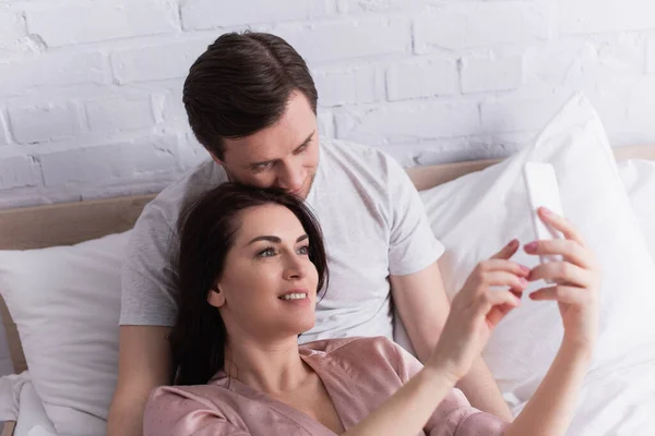 Smiling woman using cellphone on blurred foreground near husband on bed — Stock Photo