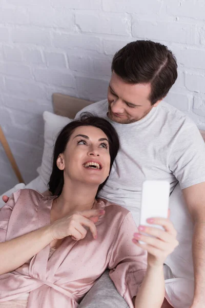 Mulher sorridente apontando para o smartphone em primeiro plano borrado perto do marido na cama — Fotografia de Stock