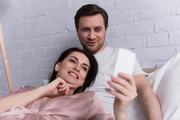 Усміхнена жінка дивиться на смартфон на розмитому передньому плані біля чоловіка в спальні — стокове фото