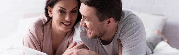 Mujer sonriente sosteniendo teléfono inteligente cerca del marido en la cama, pancarta - foto de stock