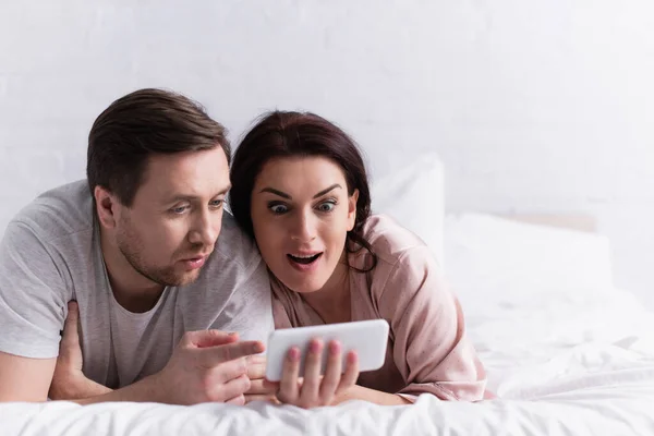 Excited adult couple looking at smartphone on blurred foreground on bed — Stock Photo